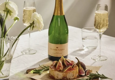 A bottle and two glasses of Mumm Napa Blanc De Noirs beside a wooden board with a Baked Brie With Apricot Compote decorated with figs