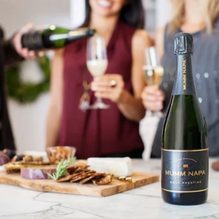 A bottle of Mumm Napa Brut Prestige sparkling wine next to a board of nibbles with people pouring wine and holding champagne glasses in the background