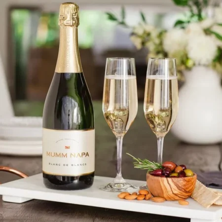 A bottle and two glasses of Mumm Napa Blanc de Blancs sparkling wine, beside two champagne glasses and a platter of nibbles including olives and almonds