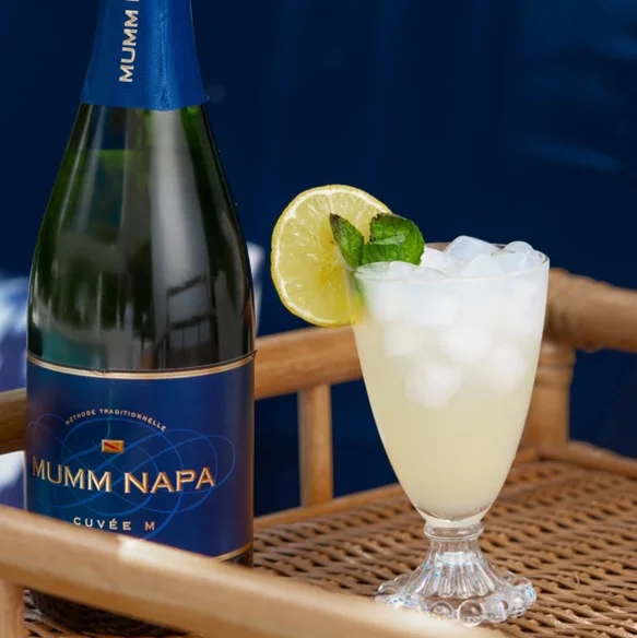 Glass of sparkling lemonade garnished with a sprig of mint and a slice of lemon next to a bottle of Mumm Sparkling Cuvée M