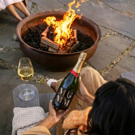 A women holds a bottle of Mumm Napa Brut Reserve sparkling wine, with a blazing fire pit in the background