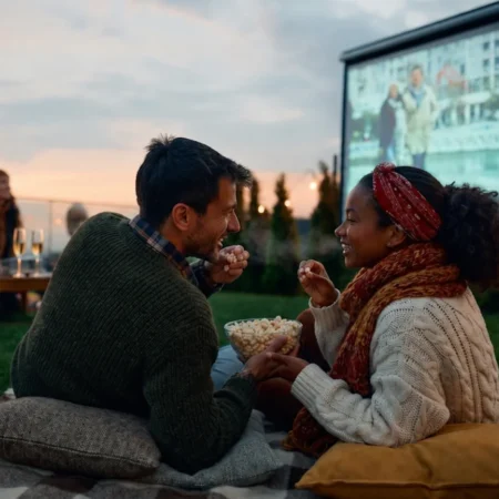 A smiling couple share a bowl of popcorn at dusk, lying on cushions with a large outdoor movie screen behind them