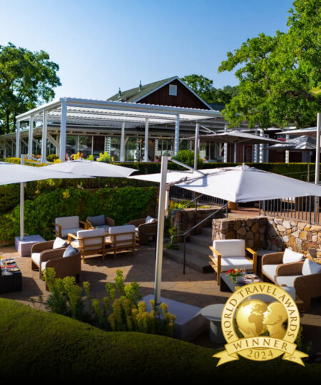 An exterior view of the Mumm Napa tasting room showing an outdoor patio and sun shades, with a superimposed golden logo that shows a figure holding a globe and the words 'world travel awards winner 2024'