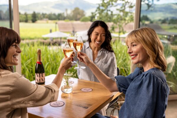 Three smiling women sitting the Mumm Napa tasting room with a rural view behind them raise their glasses of Mumm Napa sparkling wine to toast one another