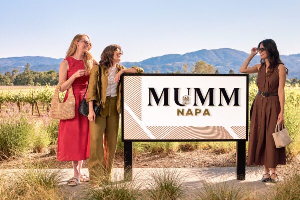 Three women stand next to an outdoor sign that says "Mumm Napa" with sunny vineyards and hills in the background