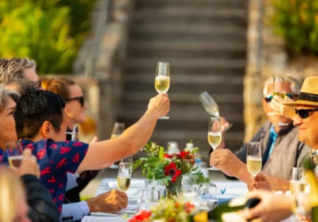 People sat on either side of a long outdoor table on a sunny day, cheers their sparkling wine glasses