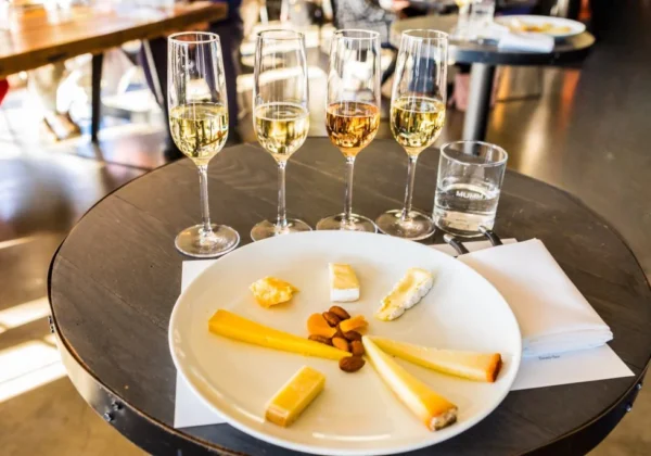 A row of four champagne flutes, each with a different sparkling wine in it, and a plate with a selection of cheeses arranged around it in front