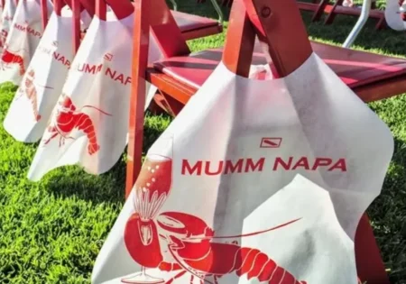 Red folding chairs with bibs hung on their backs, featuring a red lobster and Mumm Napa logo, are arranged on a green grass lawn